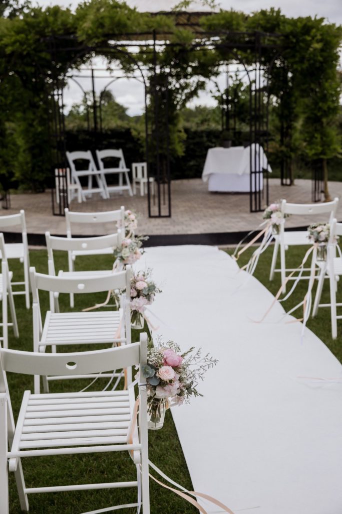 Bruiloftsopstelling buiten met witte stoelen aan weerszijden van een gangpad. Het gangpad is omzoomd met boeketten bloemen die aan stoelen zijn vastgebonden. Een met wijnranken bedekt prieel en tafel staan op de achtergrond, op een stenen platform.