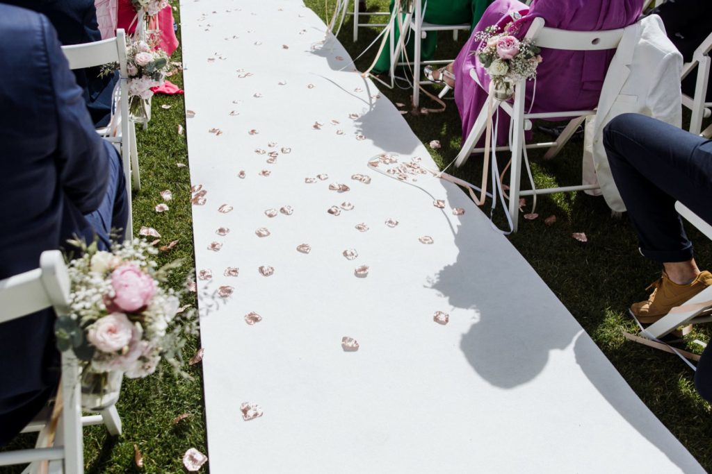 Een witte gangpadloper is versierd met verspreide roze bloemblaadjes. Witte stoelen staan langs het gangpad en zijn versierd met kleine boeketten van roze en witte bloemen. Schaduwen vallen over de loper en zonlicht verlicht de grazige omgeving.